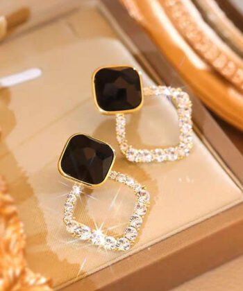 A pair of black rhombus earrings with golden frames and sparkling crystals being held on a finger