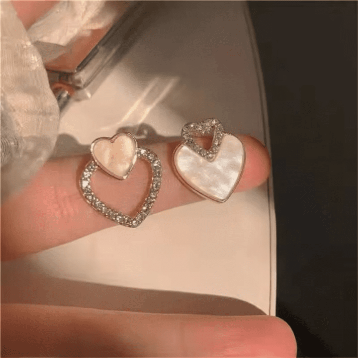 Korean-Double-Heart-Zircon-Earrings-Aish-Splash Close up of two different heart-shaped earrings with crystals and shell texture on a finger