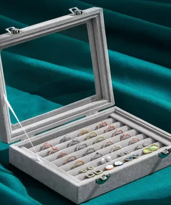 Grey velvet jewelry box with glass lid showing rings and earrings inside.
