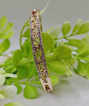 Rose gold filigree bangle with crystal accents displayed against green leaves.